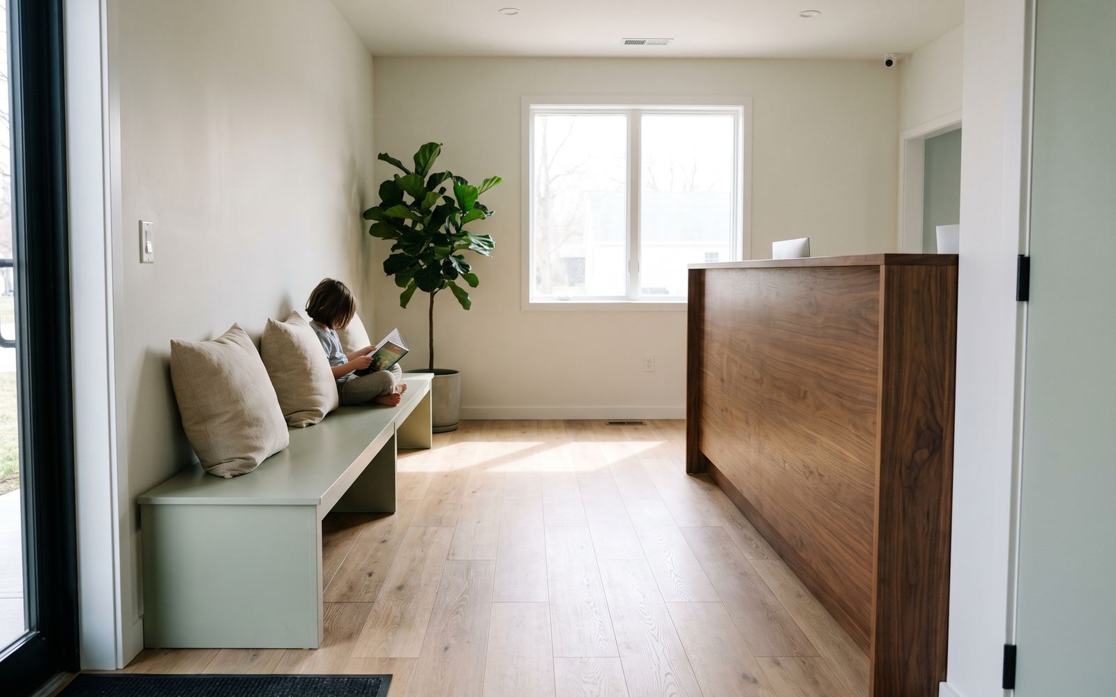 The Alder Dental Studio waiting room: a child reading on a sage-green bench beside a fiddle-leaf fig, with a walnut reception desk and morning light across the oak floor
