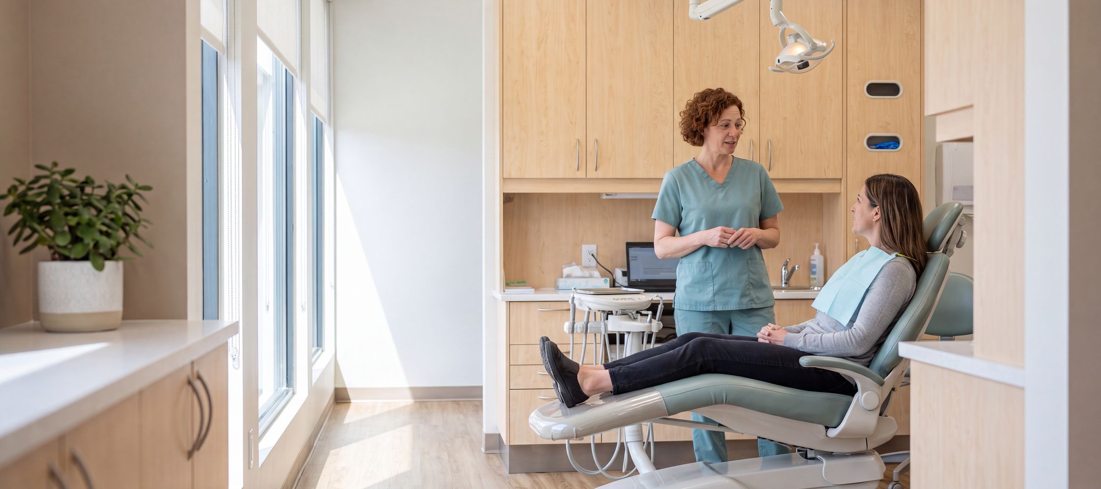 Dr. Maya Alder in teal scrubs talking with a relaxed patient reclined in a bright, sunlit dental operatory