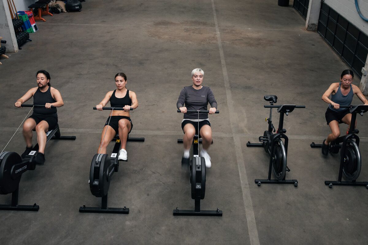 Three members mid-effort on rowing machines and air bikes in a raw industrial gym, flywheels blurred with motion