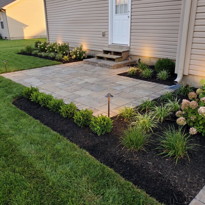 After: the same doorway with a banded stone paver patio tying into the steps, mulched beds of clipped boxwood and hydrangea, and low path lighting