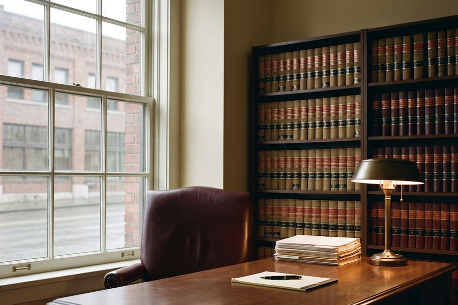 A Harbor & Vale office — a walnut desk and oxblood leather chair beside a tall window looking onto a downtown brick facade, with shelves of bound legal reporters.