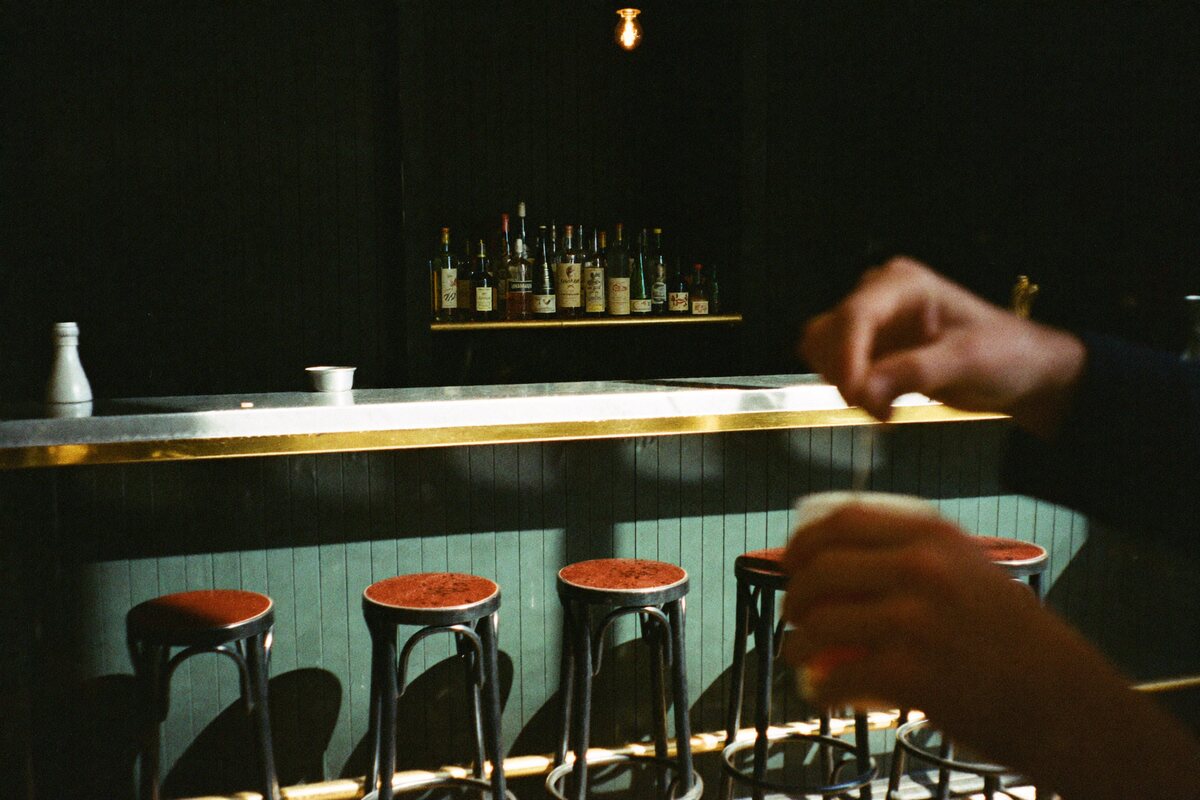The Pilcrow bar during service: a zinc counter, backbar bottles lit from below, and a bartender stirring a drink in the foreground