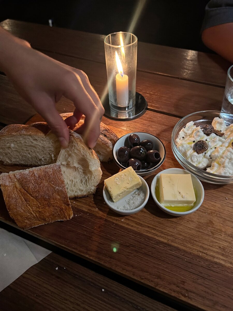 A shared starter board on dark oak: torn wood-fired sourdough, cultured butter, olives and whipped ricotta, with a hand reaching in to tear bread