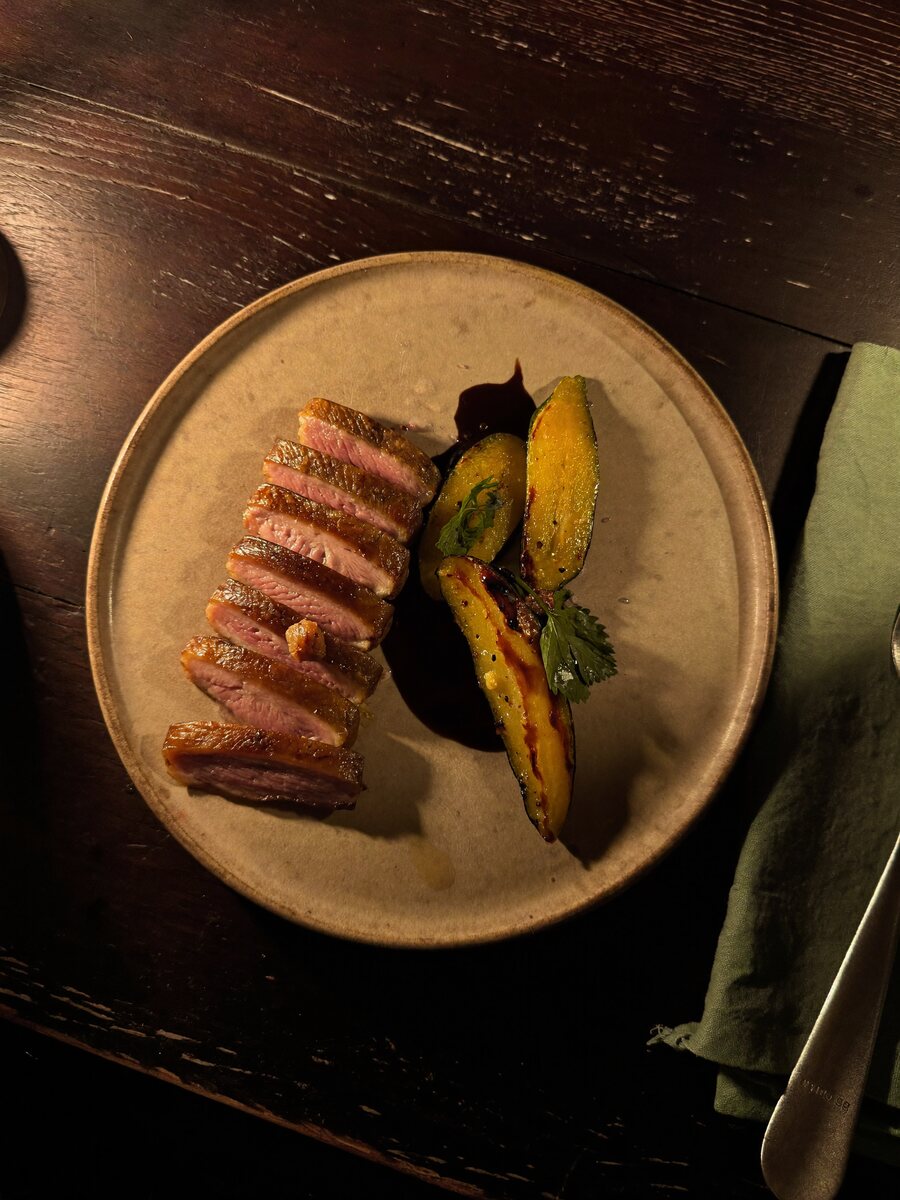 Seared duck breast sliced and fanned on a stoneware plate with roasted squash, charred scallion and a dark reduction, lit by candlelight