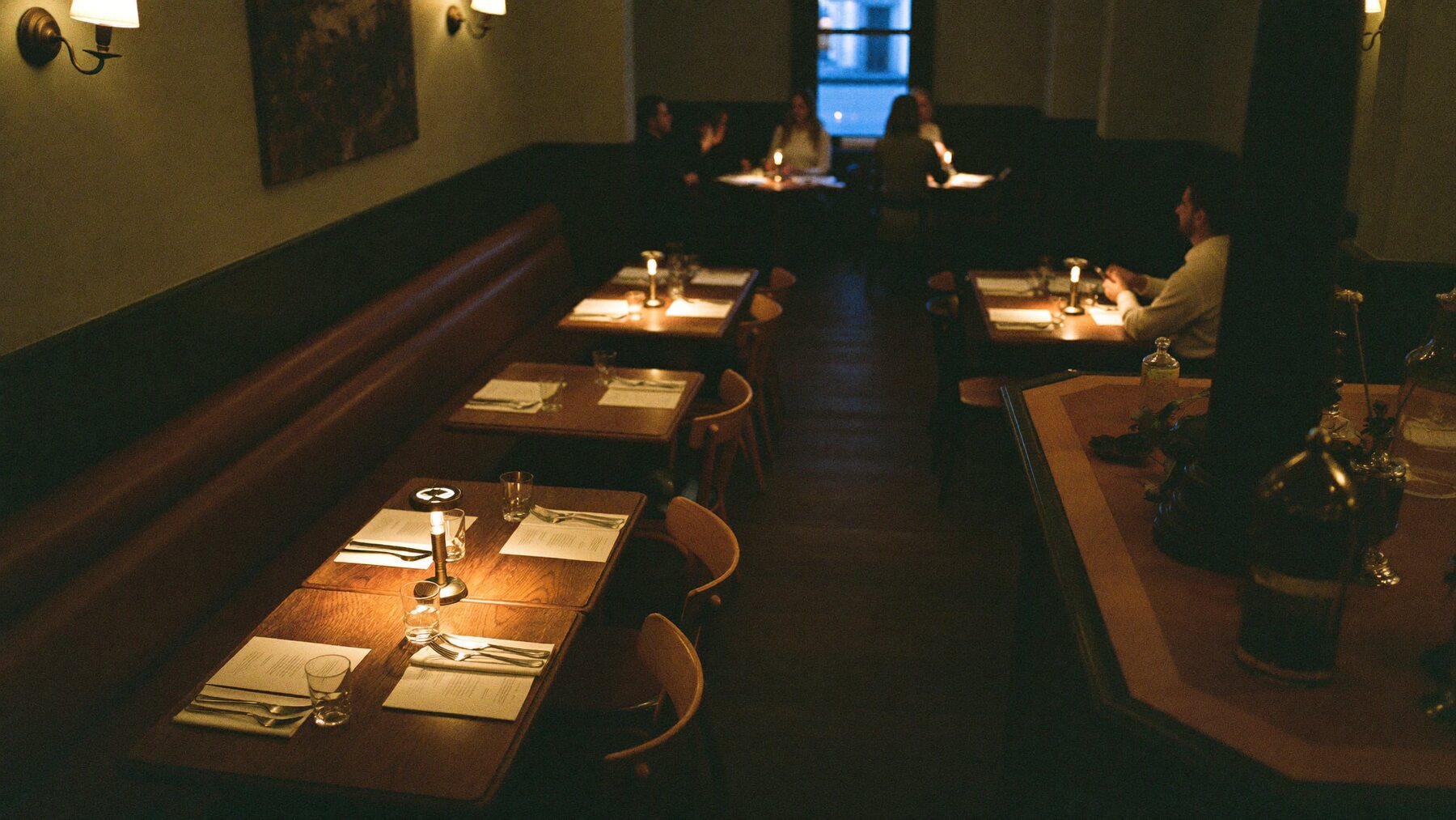The Pilcrow dining room at the start of dinner service: worn oak tables set with paper menus and lit brass candle lamps, a leather banquette down one wall, warm pools of low light