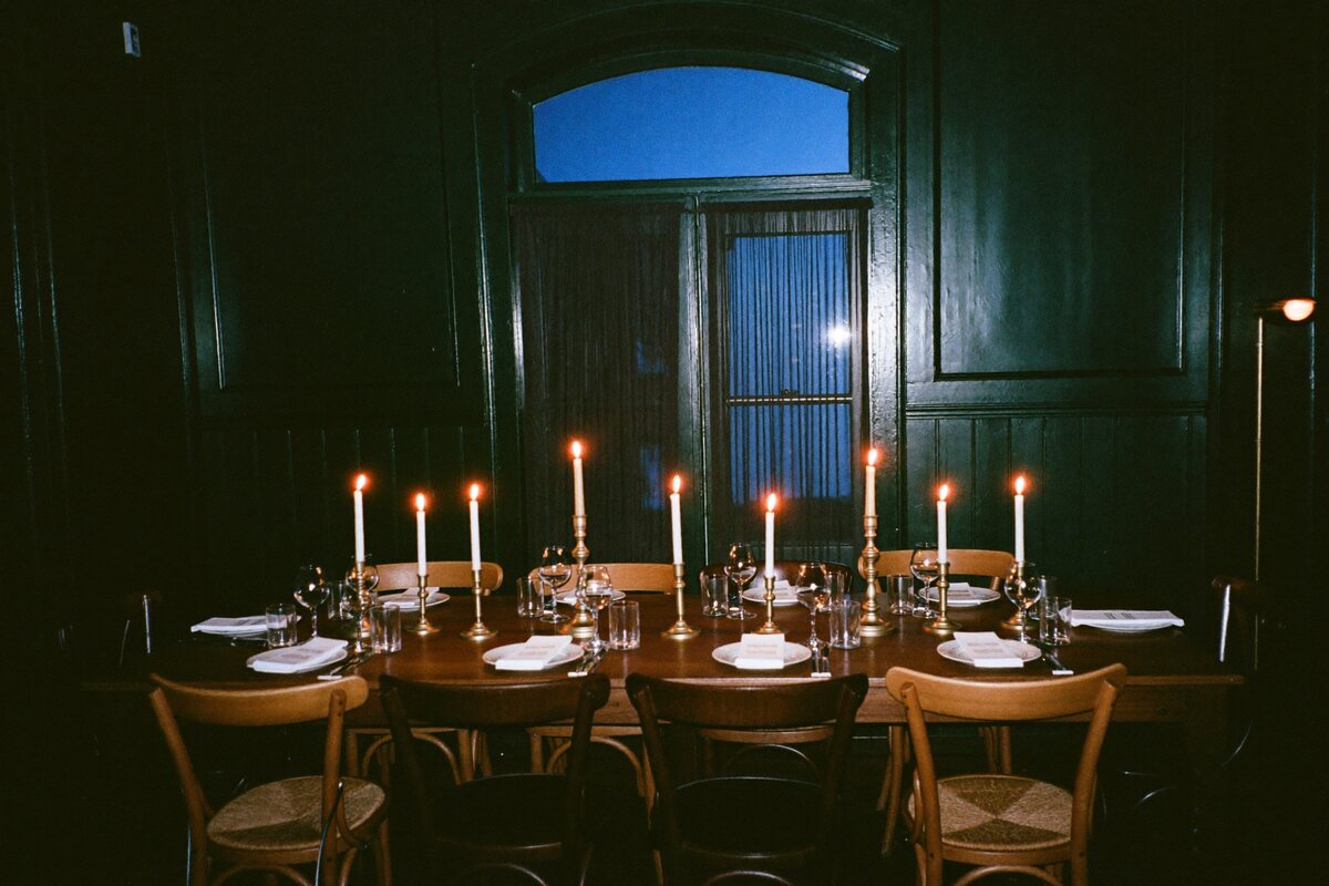 The Pilcrow back room set for twelve: one long oak table down the middle, mismatched chairs, brass candle lamps and taper candles lit end to end, dark panelled walls