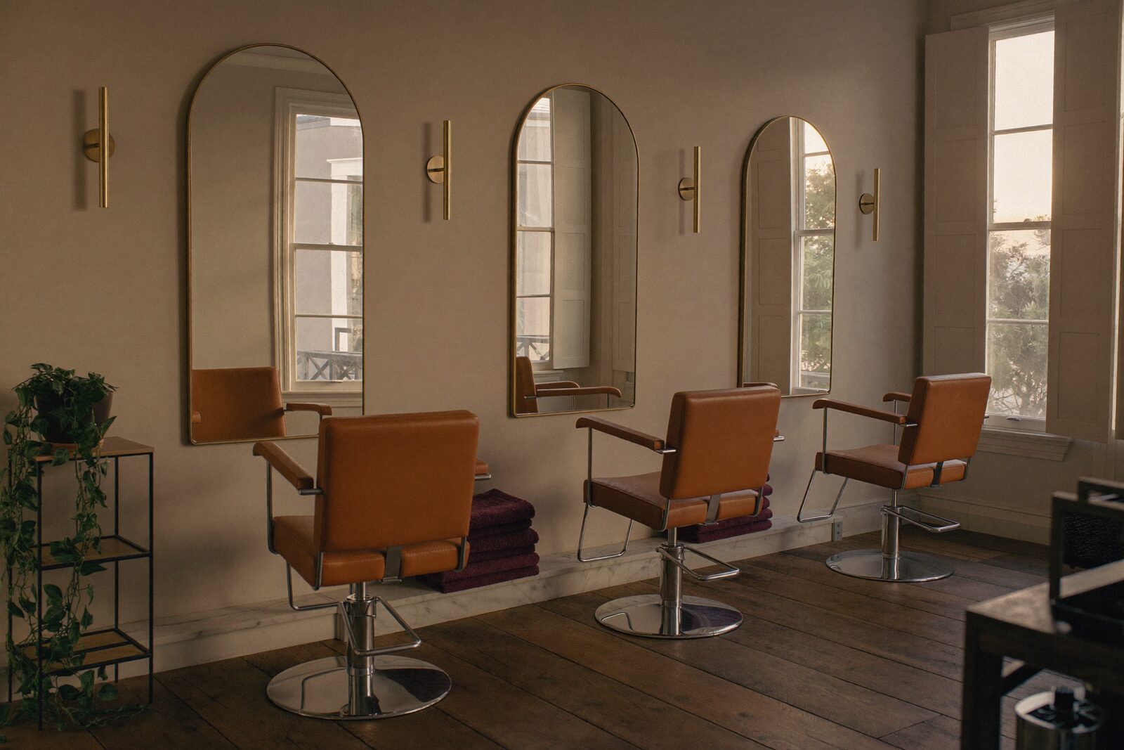 The Marcel Avenue Studio floor: three styling chairs along a bone plaster wall, each under a tall arched brass-framed mirror, with a stack of mulberry towels on a marble ledge