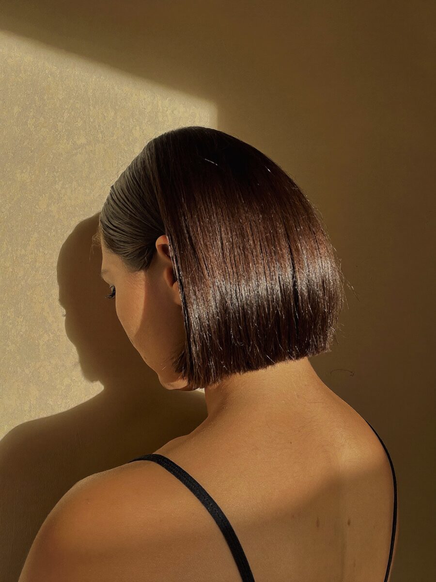 A fresh blunt collarbone-length cut in a cool brunette, photographed from behind against a plaster wall